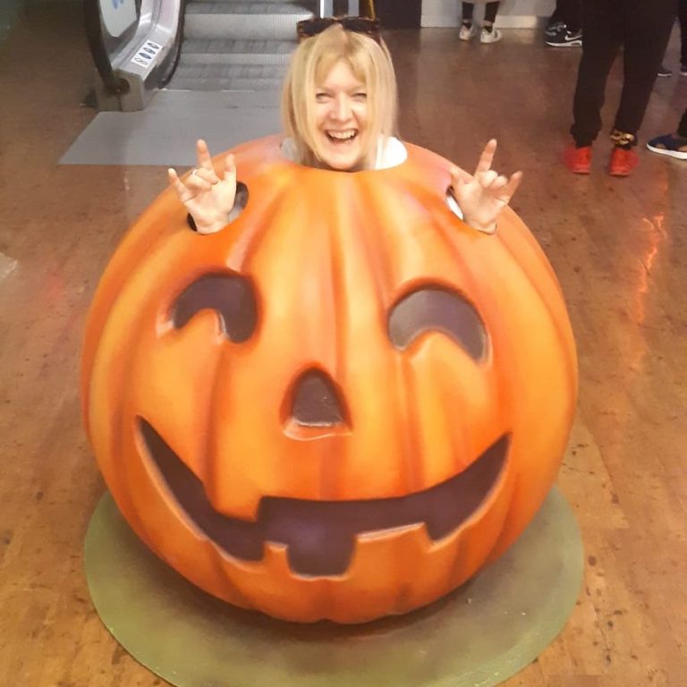 Person smiling and making rock hands from inside a large, cheerful pumpkin decoration.