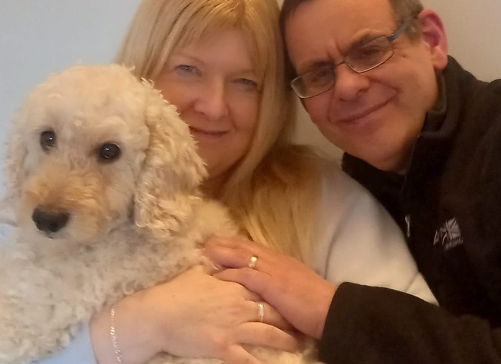 A woman and a man smile while holding a fluffy white dog between them.