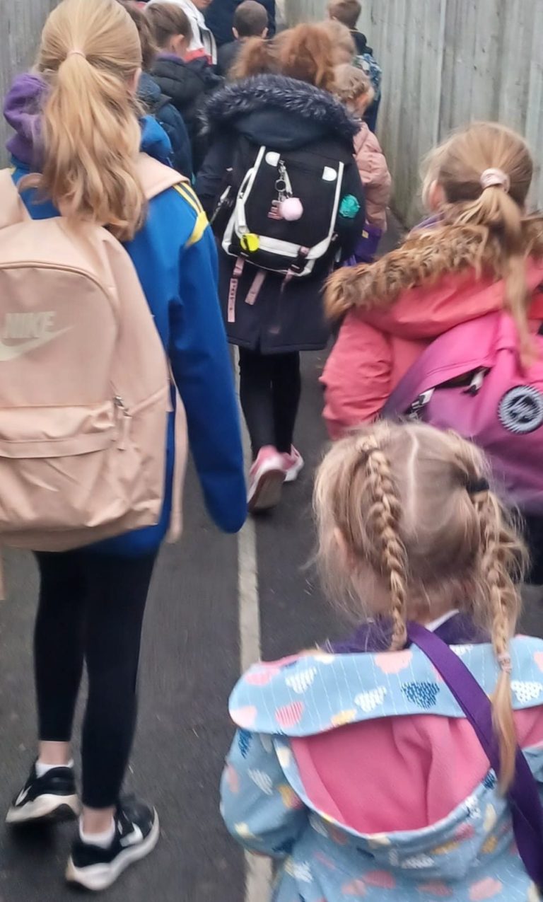 A group of children walking along a narrow path, wearing backpacks.